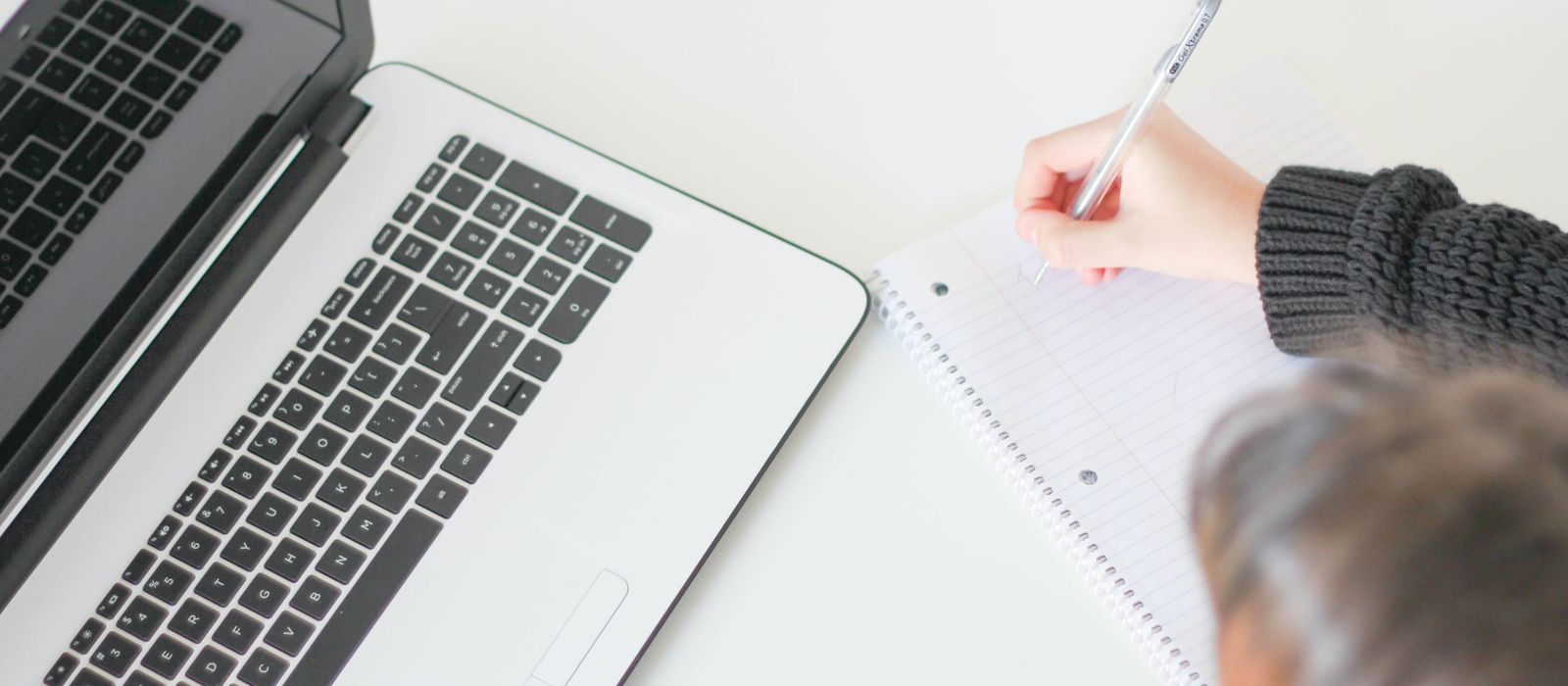 A writer taking notes in a ruled notebook beside a laptop on a bright desk